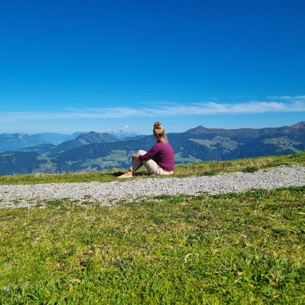 Sonja Schüssele Frau sitzt auf einem Bergweg und blickt auf eine malerische Landschaft.