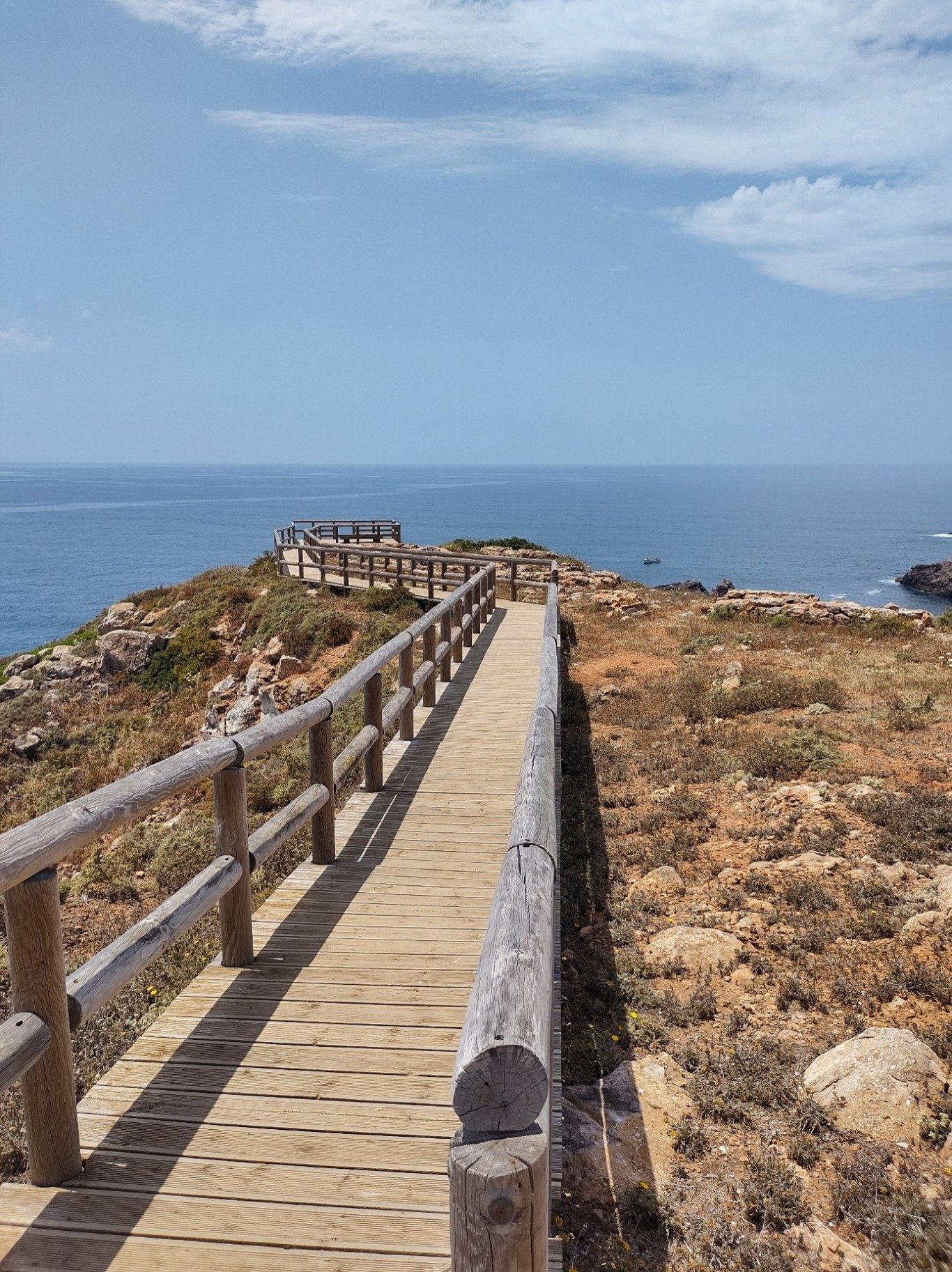 Holzsteg führt über eine felsige Küstenlandschaft zum Meer unter blauem Himmel.